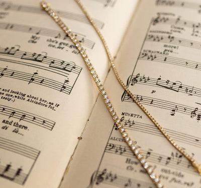 Two Essential Tennis Bracelet, which are classic tennis bracelet with diamonds set in 18kt yellow gold on a music sheet. 